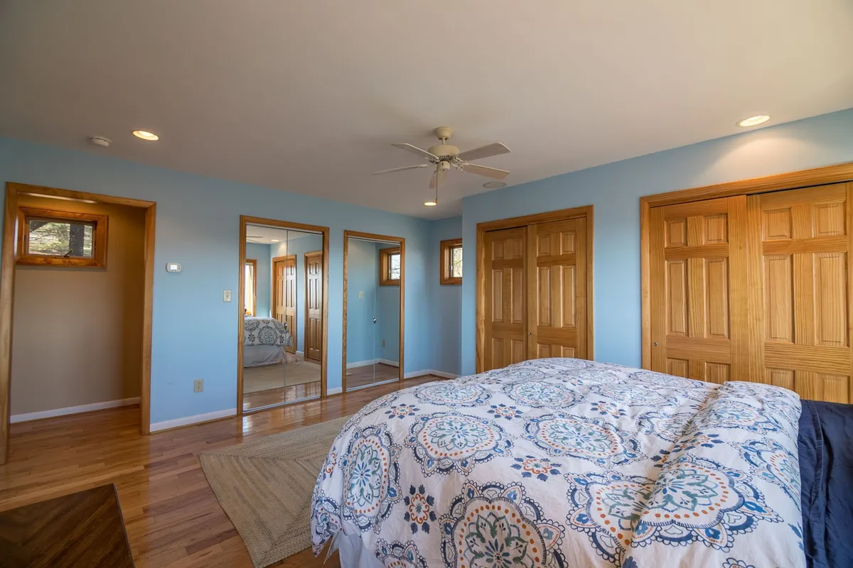 A tranquil bedroom scene shows a bed with patterned bedding positioned to receive natural light from nearby windows. Wooden doors and a mirrored closet add to the room’s sense of space and calm, evoking a restful retreat after active outdoor days.