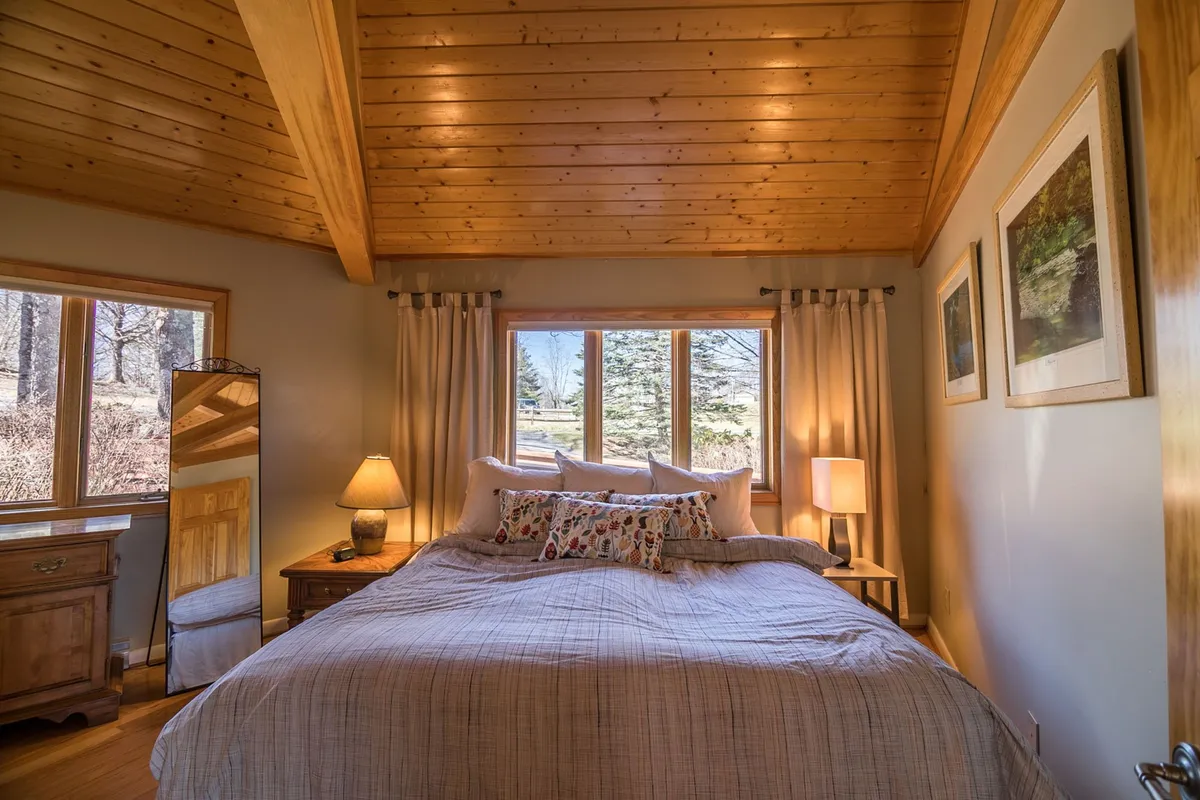 A serene bedroom with a plush bed, decorative pillows, and a wooden ceiling, illuminated by soft light streaming through large windows. The placement of the bed invites guests to wake up to beautiful winter vistas or unwind in peace after a day of snowy adventure.