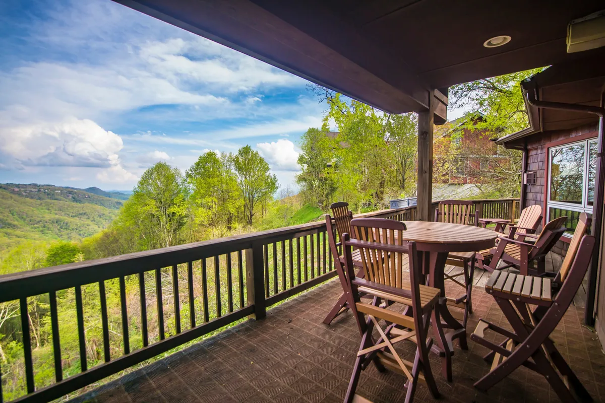 The image features a serene wooden deck set with comfortable chairs and a round table, all overlooking breathtaking, layered mountain views framed by lush greenery. The inviting outdoor space suggests moments of dining, relaxation, and taking in sunrises or sunsets against the backdrop of Blowing Rock's natural beauty.