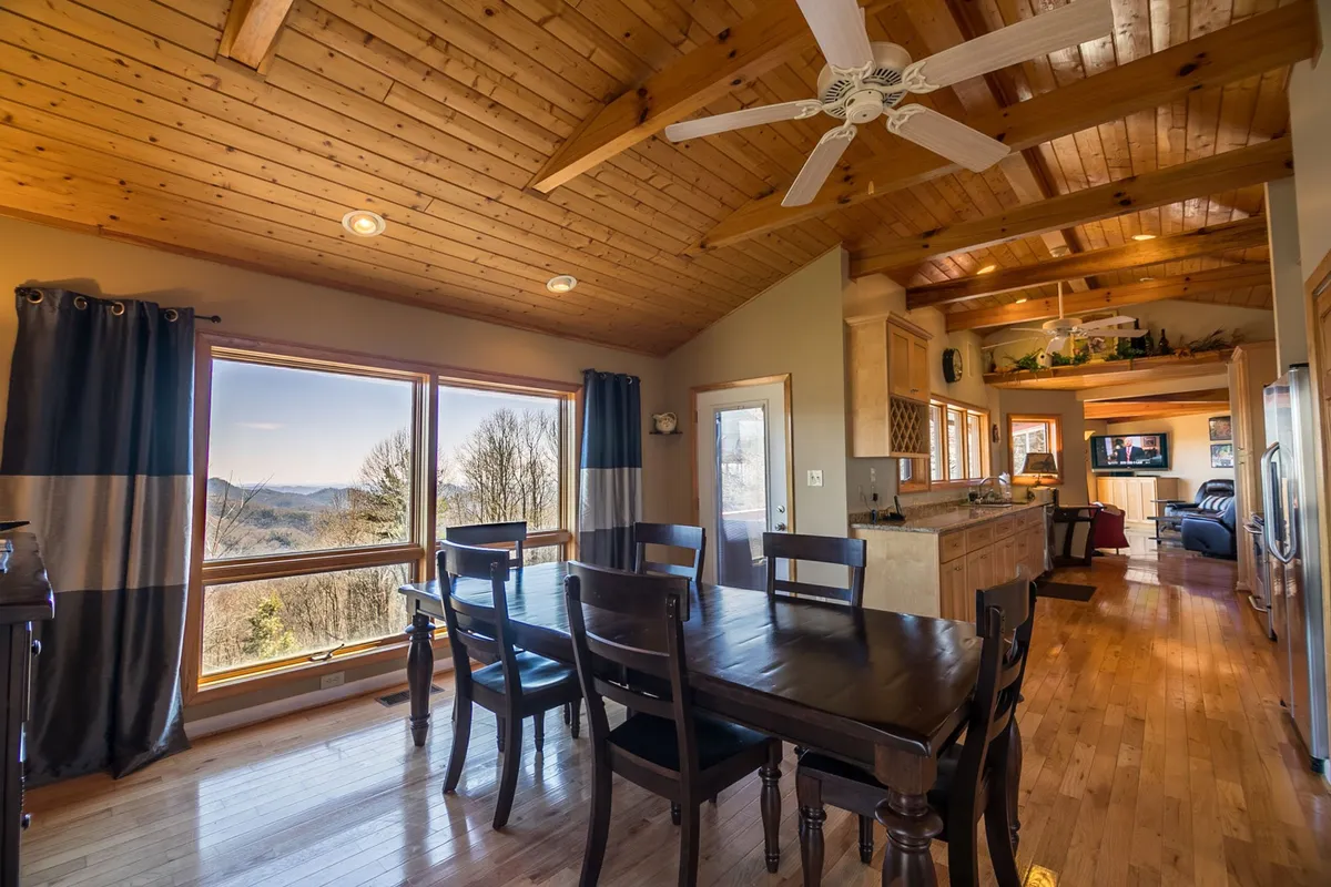 A wide view of the open dining area with a large dining table, panoramic windows revealing stunning mountain views, exposed wooden ceiling beams, and a glimpse into the adjacent kitchen area. The space feels warm and perfect for group meals or intimate dinners.