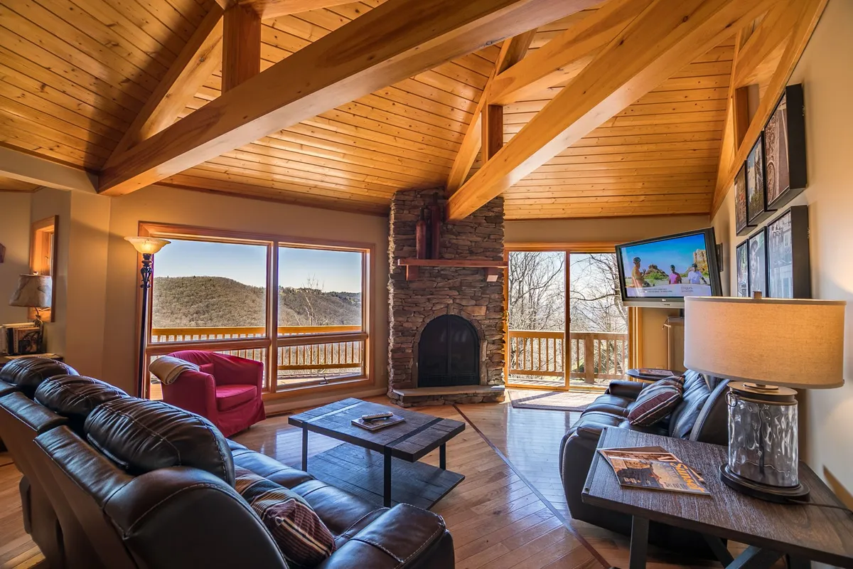 This image captures the heart of the round house living room featuring soaring wooden beams, a striking stone fireplace, comfortable leather sofas, and expansive windows framing the mountain landscape. The natural light and cozy furnishings invite relaxation and socializing.