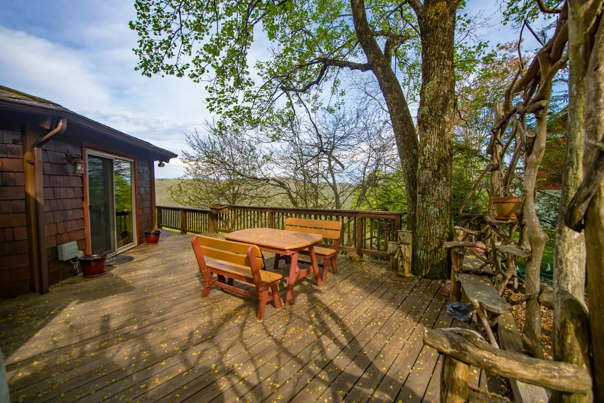 A stylish wooden deck surrounded by trees, featuring a dining table ideal for gatherings. The panoramic views and filtered sunlight through the leaves create an inviting, serene atmosphere, highlighting the natural connection between indoor and outdoor spaces.