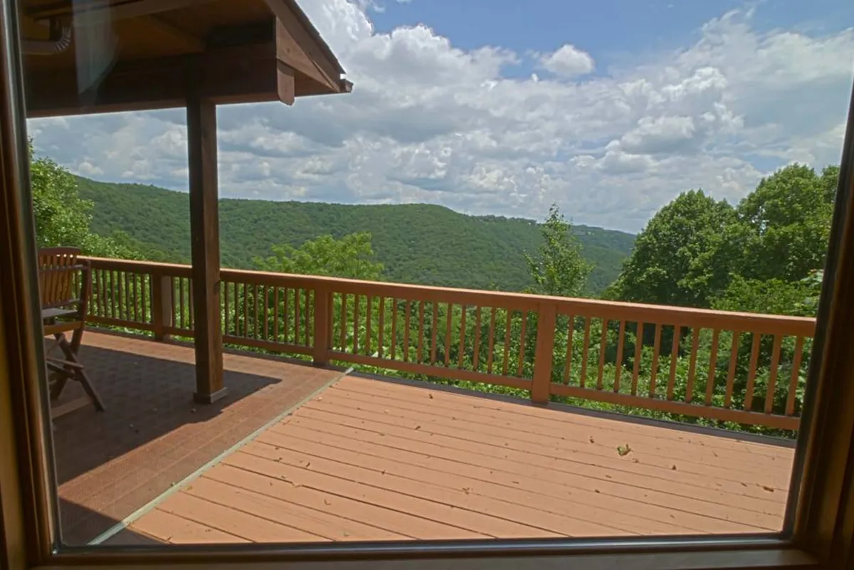 A spacious wooden deck offers sweeping mountain views under a dramatic cloudy sky. The deck is framed by rustic railings and surrounded by lush natural landscape, inviting guests to relax, gather, or simply soak in the panorama. The elevation allows for unobstructed vistas of the rolling ridges and seasonal changes in the landscape.