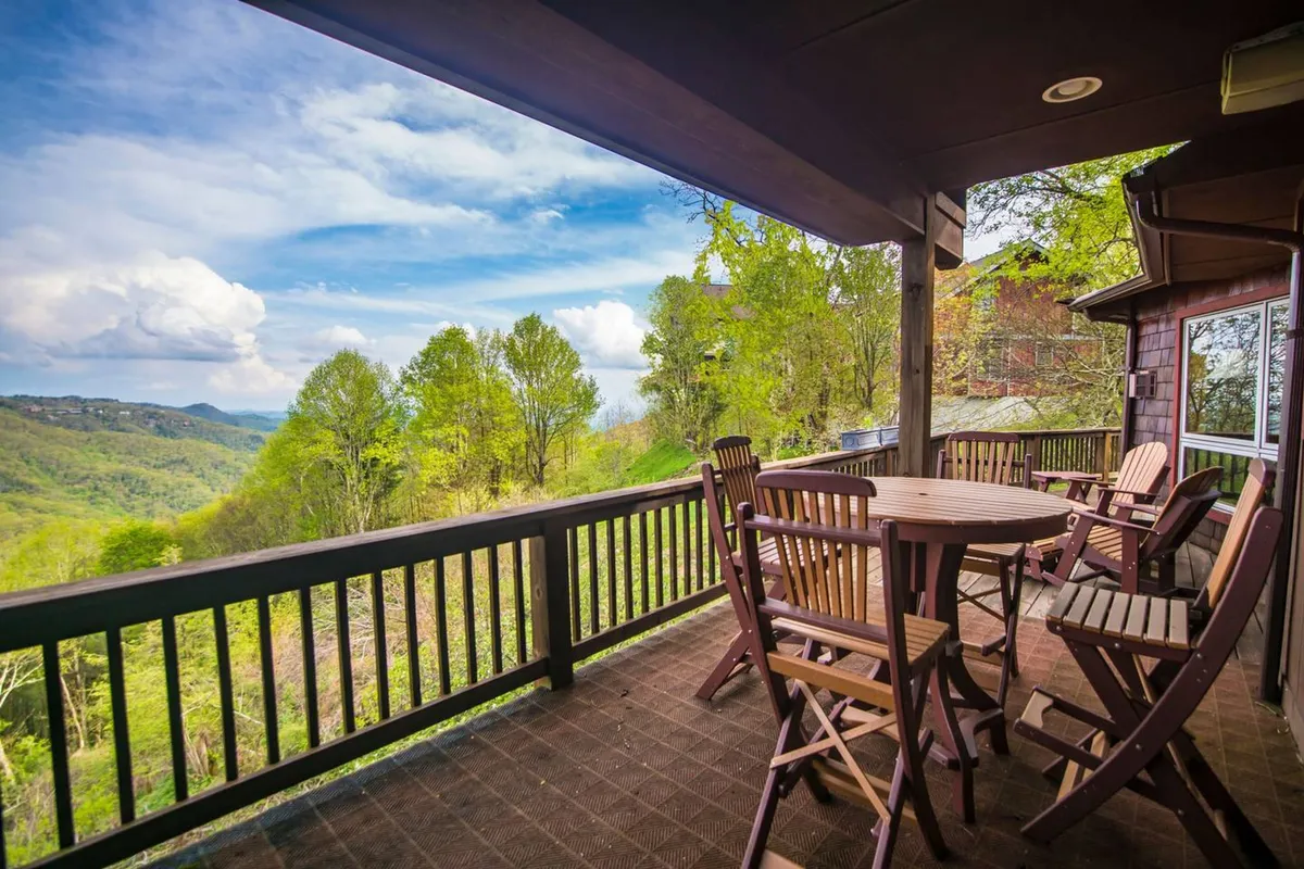 A spacious wooden deck with a table and chairs, set against a breathtaking mountain backdrop. The area is bathed in natural light, evoking a sense of peace and inviting guests to linger outdoors. The deck is perched above the landscape, offering unobstructed views and a seamless transition from inside to outside.
