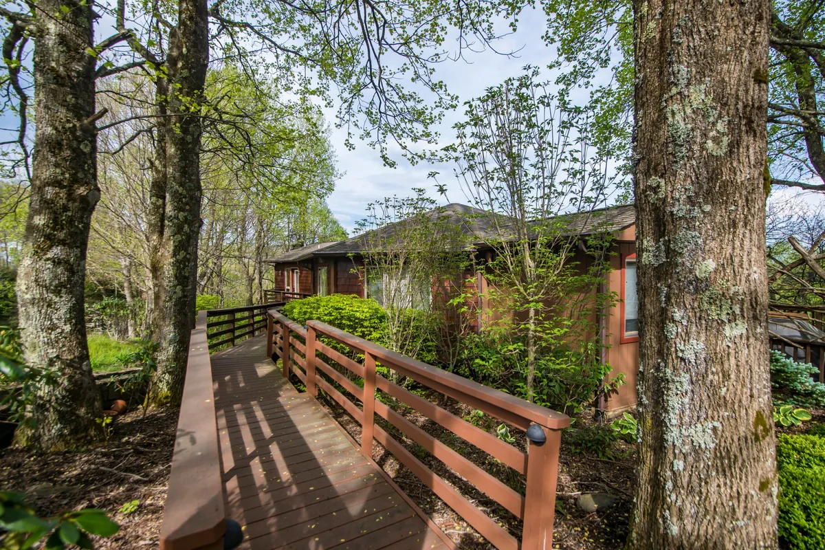 A tranquil wooden walkway meanders through lush greenery, leading to the house’s facade nestled among tall trees. The scene suggests a secluded retreat and invites exploration of the natural surroundings, embodying the peacefulness and adventure of a mountain escape.