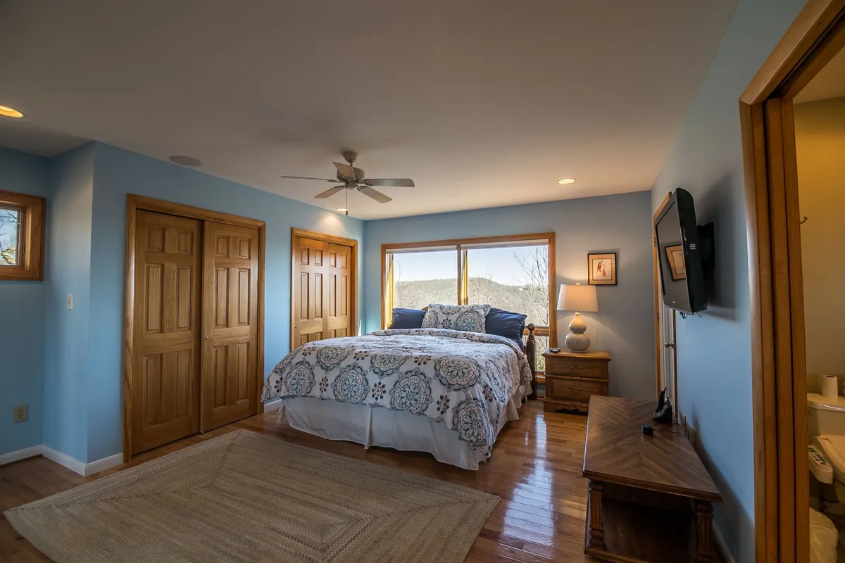 A cozy bedroom with a comfortable bed, windows inviting in daylight, a wardrobe, nightstand, and a lamp for evening relaxation. The tranquil color palette enhances the restful atmosphere.