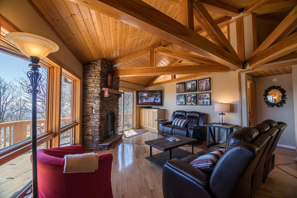 A warm and inviting living room featuring a striking stone fireplace as the centerpiece, flanked by large windows that let in natural light and offer glimpses of the snow-dusted landscape outside. Comfortable seating surrounds the fireplace, inviting guests to gather, relax, or enjoy a cozy movie night.