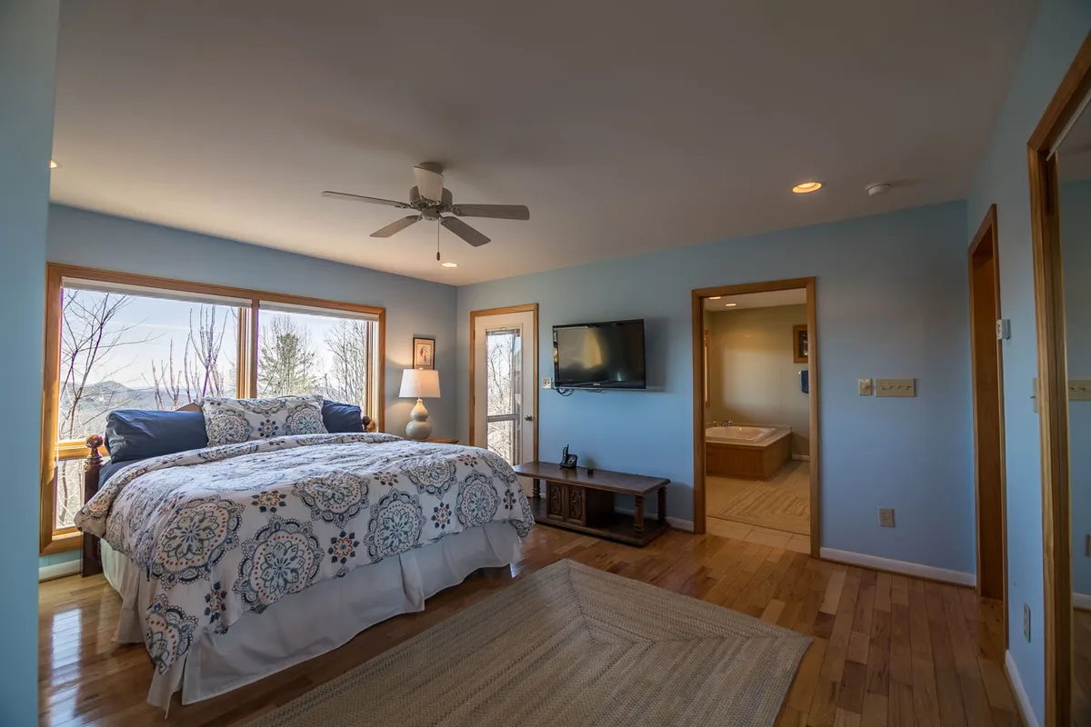 A spacious bedroom featuring a large bed with decorative bedding, floor-to-ceiling windows that flood the room with natural light, and wooden furniture that adds a touch of warmth. The serene ambiance invites relaxation and offers beautiful views right from the bed.