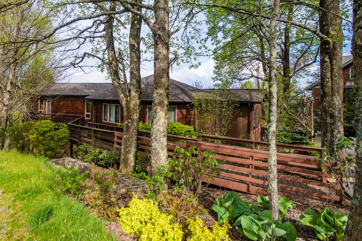 The house's exterior is framed by lush greenery and decorative fencing, with a visible level grassy area ideal for pets and play. The wooden structure hints at the welcoming deck above, while the setting radiates a tranquil, pet-friendly environment.